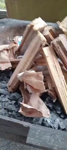 This media file captures a close-up view of a fire being built in what appears to be an outdoor setting, possibly a barbecue pit or fireplace. The primary subjects are several pieces of firewood, stacked in an A-frame structure over a bed of charcoal. Underneath the wood, crumpled brown paper is being used as tinder, with visible flames igniting it.

The activity taking place is the process of starting a fire. The paper is actively burning, producing flames and some smoke, and the heat from the fire is beginning to affect the paper, causing it to curl and darken. The wood itself is not yet burning significantly, but it is positioned to catch fire as the flames grow.

Notable details include the rough texture of the wood, some of which shows signs of being split. The charcoal below is dark and appears to be partially glowing. The lighting suggests it might be daytime, though the specific time of day is not evident. There are no visible signs of weather conditions. The setting appears to be functional, designed for burning materials. No people are visible in the frame, and there are no discernible emotions conveyed due to the absence of human subjects.

No visible text is present in the media file.