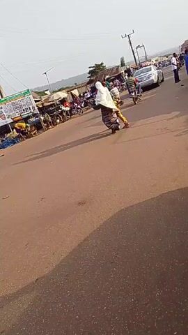 The video shows a busy street scene in what appears to be a town or city in Africa. The road is paved and mostly reddish-brown in color. There are people walking, riding motorcycles, and standing around. Some people are wearing traditional clothing, while others are dressed in more modern attire. Several motorcycles are visible on the street, along with at least one car. There are shops and buildings lining the street, and some street vendors are selling goods. The weather appears to be sunny and clear. The camera seems to be positioned inside a vehicle.
