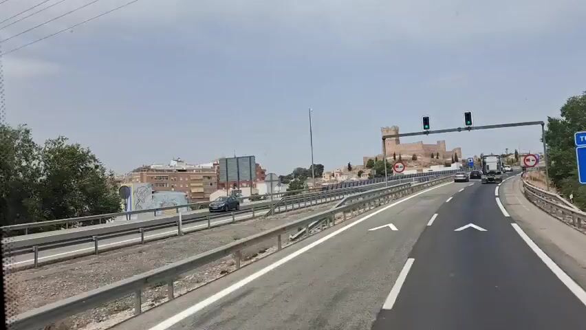 A forward-facing, first-person perspective shot from a vehicle traveling on a highway. The highway has multiple lanes, with guardrails on both sides. The road is paved and marked with white lane lines and directional arrows. In the distance, on a hilltop, stands a large, ancient-looking stone castle with several towers and walls. The sky is overcast. Traffic signs are visible, including traffic lights, a red circular sign with a horizontal bar, and a blue rectangular sign indicating a tunnel. Several vehicles, including cars and a van, are also visible on the highway. Buildings, including a red apartment building, are visible to the left of the highway. Trees are scattered throughout the landscape.