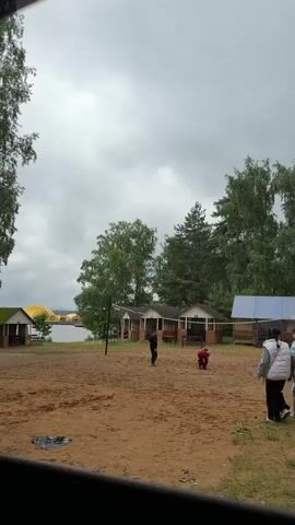 The video shows a sandy area with several small structures in the background, possibly cabins or shelters. A lake or large body of water is visible to the left, surrounded by trees. The sky is overcast with clouds. In the foreground, four people are walking across the sand. Two of them, appearing to be young women, walk together towards the right side of the frame. Another person, dressed in dark clothing, walks towards the left. A fourth person, wearing a red jacket, is crouched down in the middle distance, possibly playing a game. A volleyball net is visible in the background. The scene appears to be a recreational area.