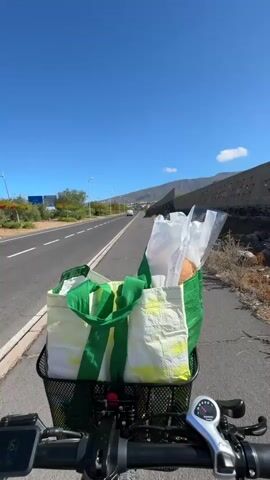 The video shows a first-person perspective of someone riding an electric bicycle on a paved road with a basket full of groceries. The basket contains at least two large reusable shopping bags, one green and white, and another white with yellow accents. Inside the bags, some items are visible, including a loaf of bread wrapped in plastic.

The setting is outdoors on a bright, sunny day with a clear blue sky and a few wispy clouds. The road appears to be a well-maintained asphalt surface, with a white line marking the edge of the bike lane or shoulder. To the left of the road, there is a roadside embankment with some vegetation. To the right, a low stone wall runs parallel to the road, and beyond it, dry, scrubby vegetation can be seen. In the distance, hills or mountains are visible, with buildings and streetlights lining the road. The overall impression is a warm, dry climate.

The activity is bicycle riding, likely for shopping or returning from a shopping trip. The rider is on a road, suggesting a journey through a town or along a scenic route. The presence of groceries implies a mundane, everyday activity. The time of day appears to be midday, given the bright sunlight and clear sky.

Notable details include the electronic display on the handlebars of the bicycle, which shows a speed of 19 km/h and a battery level of 65%. The handle grips and brake levers are also visible. The overall mood is peaceful and perhaps a little adventurous, with the rider enjoying a pleasant ride in good weather. The location cues (stone walls, dry vegetation, distant hills) align with the context of Adeje, Spain, which is known for its sunny climate and varied landscapes.