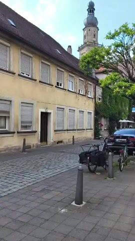 The video shows a street scene in what appears to be a European town. On the left is a pale yellow building with a dark tiled roof and numerous windows, most of which are covered by roller shutters. The street in front of the building is paved with cobblestones. To the right, a darker building is partially visible, covered in ivy, with a tall clock tower rising above it. A large green tree is in the foreground on the right, obscuring part of the view. Parked on the right side of the street are two bicycles and a dark-colored car. The sky is blue with some white clouds. There are no people visible in the scene. The overall impression is one of a quiet, historic street.