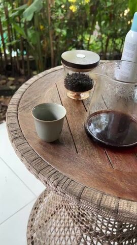 A close-up shot shows a person pouring dark liquid from a clear glass pitcher into a small gray teacup on a round, weathered wooden table with a woven base. The table also holds a small glass jar with a metal lid containing what appears to be loose tea leaves. The background consists of green plants and trees, suggesting an outdoor or garden setting. The overall lighting is soft and natural, highlighting the textures of the table and the tea.