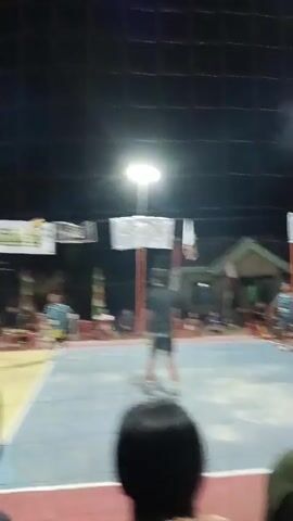 A volleyball game is being played at night on an outdoor court. The court is lit by overhead lights. There is a net dividing the court into two sides. On one side of the net, there are several players in blue uniforms. On the other side of the net, there are players in maroon and blue uniforms. The ball is in the air, and players are actively engaged in the game. Spectators are seated in the foreground, watching the match. Banners are hanging above the court.
