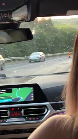 This is a first-person perspective video shot from inside a moving car. The camera is focused on the car's dashboard and the road ahead. The dashboard has a large infotainment screen displaying a navigation map, as well as climate controls with digital temperature readouts. A person's shoulder and hair are visible in the bottom right corner of the frame, indicating the videographer is a passenger. Outside the car, a road is visible with trees and hills in the background. There are other cars on the road, and traffic cones are present. The sun appears to be setting, casting a warm light.