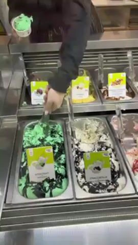 Here is a description of the video.
A person in a black top and a striped apron is scooping green ice cream into a small white paper cup, from a stainless steel container labeled "After Eight Bites". The stainless steel container is one of a series of similar containers holding different flavored ice creams. Each container has a label with the name of the flavor. The containers are refrigerated behind a glass window. Above the glass is a shelf with miscellaneous items, including a sign. The ice cream flavors visible include chocolate cherry, lemon, hazelnut, stracciatella, and a berry flavor. The person hands the cup to someone standing in front of the counter.