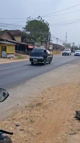 A black pickup truck drives on the left side of a paved road. Several cars are driving on the right side of the road, going in the same direction as the truck. A white SUV is in the lead on the right, followed by a white sedan, and then another white sedan. The road is lined with buildings and trees on the left and dirt and vegetation on the right. Overhead power lines crisscross the sky.