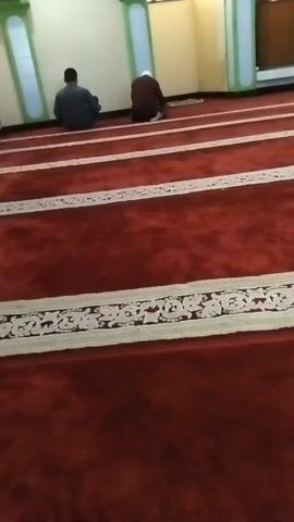 A wide shot of a mosque interior shows a red carpet with white striped borders. Several people are visible. In the foreground, the carpet fills the frame. Further back, two men are seen kneeling on the carpet, facing away from the camera. To their right, another person is also kneeling. In the background, near an open doorway, two men are standing, and a child is sitting on the carpet. The walls are a light color with green accents and tall, narrow windows.