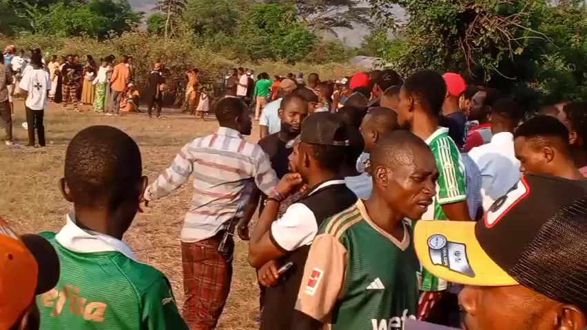 The video shows a wide shot of a large outdoor gathering of people in a dry, grassy field. In the background, there are trees and hills, with some buildings visible on the right side. The sky is clear and bright, suggesting it is daytime. A crowd of men, women, and children are scattered across the field, some standing, some walking, and some appearing to be talking to each other. Several people are wearing colorful clothing, and some appear to be wearing uniforms or team jerseys in green and white. The overall atmosphere seems to be one of a community event or gathering.