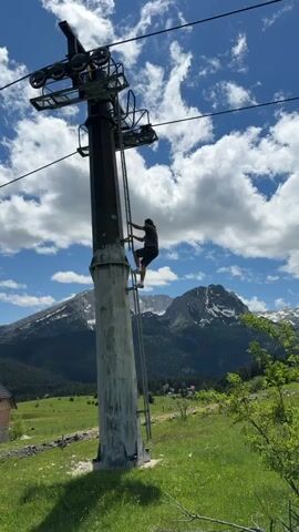 The video features a man climbing a metal ladder attached to a tall, dark grey metal pole, likely a ski lift tower. He is wearing a grey t-shirt, dark shorts, and light-colored sneakers. As he climbs, he reaches a certain height, then turns to face the camera, smiling and briefly posing with one arm outstretched.

The scene is set outdoors on a bright, sunny day with a clear blue sky dotted with fluffy white clouds. In the background, majestic mountains with patches of snow are visible, stretching across the horizon. The foreground and midground consist of a vibrant green grassy field, suggesting a warm season. A small building with a wooden facade is partially visible on the left side of the frame in the initial shots. The overall impression is one of leisure and adventure in a scenic mountainous environment, specifically in Pitomine, Montenegro.