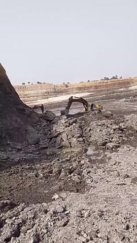 The video shows an open-pit mining operation with two excavators actively working. The foreground is filled with grey rock and mud. In the background, there are layered terraces of earth and some vegetation is visible on the horizon. One excavator is actively digging into a pile of rocks. The other excavator is positioned further away and is also working on the terrain. A person is also visible in the distance near one of the excavators. The sky is a pale blue and the lighting suggests it is a bright, sunny day.