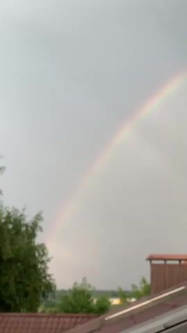 A rainbow arcs across a cloudy sky. The lower portion of the image shows the roof of a building, with trees and distant structures visible. The camera angle is looking slightly upwards towards the sky.
