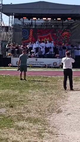The video shows an outdoor stage with a large LED screen displaying a colorful, geometric pattern. On stage, a group of people in traditional white and black clothing are performing, some playing instruments. Two men stand in the foreground, facing the stage; one is holding a camera. A woman in a red and white dress and a white hat is visible on the right. The ground is covered in grass and dirt, and the sky is clear and blue. There's a banner on the stage that appears to be in Romanian. The overall impression is of an outdoor cultural event.