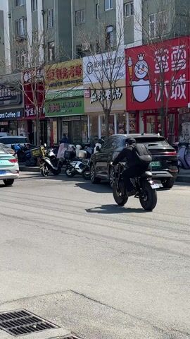 The video shows a street scene in what appears to be a busy commercial area. Various shops and businesses line the street, with signs in Chinese visible. Several cars and motorcycles are parked or moving along the street. In the first shot, a person on a motorcycle drives away from the camera. In the second shot, a person wearing a helmet and mask rides a scooter toward the camera. The final shot shows the motorcycle from the first scene driving further down the street. The overall impression is of a typical urban street scene in China.
