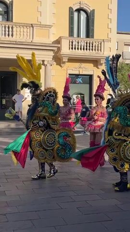 This video captures a vibrant street performance likely during a festival or parade in Sant Cugat del Vallès, Spain. The main subjects are individuals dressed in elaborate, colorful costumes, reminiscent of carnival or traditional dance attire. These costumes are characterized by large, ornate headdresses adorned with feathers in shades of yellow, pink, and blue. The performers also wear sequined and fringed outfits in bright colors, including pink skirts and boots, and elaborate masks or face coverings that contribute to their theatrical appearance.

The scene is set on a paved street, with a light-colored, multi-story building forming the backdrop. The architecture of the building, with its balconies and arches, suggests a European setting. The ground is tiled in a pattern of grey and brown squares. In the background, other people are visible, some watching the performance, while others walk by.

The activity taking place is a procession or dance performance. The individuals in costume are moving and gesturing, suggesting they are part of a coordinated display. The overall mood appears festive and celebratory, amplified by the bright costumes and energetic movements. The time of day appears to be daytime, possibly late afternoon, given the warm lighting and shadows cast on the ground, indicating sunny weather.

There is a visible sign on the building in the background with a logo that appears to be "CaixaBank", a Spanish bank. The flags attached to some of the costumes feature colors like red, yellow, and green, possibly representing a national or regional flag, or symbolic colors for the performance. The detailed craftsmanship of the costumes, especially the intricate gold and turquoise sequined embellishments, are a notable detail.