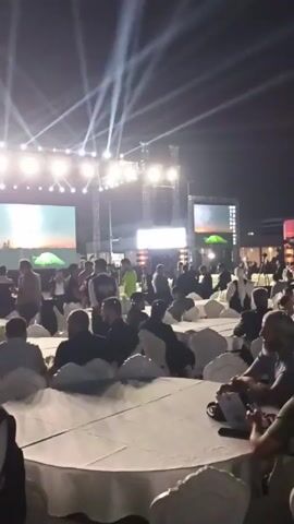 An event unfolds outdoors at night, illuminated by spotlights and large screens. The audience sits at round tables, facing a stage where a video displays a scene with tents. People are milling about, and some seem to be engaged in conversation. The atmosphere appears formal and organized, with a focus on visual presentation.