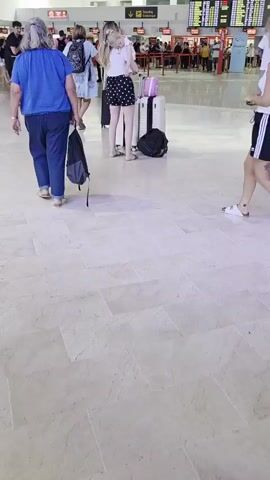In an airport terminal, a woman with blonde hair and a white tank top and black shorts stands facing away from the camera, holding a baby. She has tattoos on her right leg and left arm. Next to her are two white suitcases and a black backpack. To her left, a woman with gray hair, wearing a blue t-shirt and jeans, walks away from the camera, pulling a black bag with wheels. Further back, more people and luggage are visible, along with airport check-in counters and departure boards. The floor is tiled in a light color.