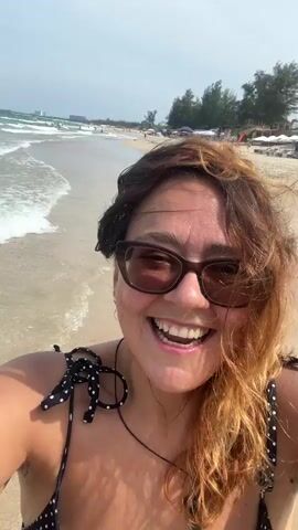 A woman stands on a sandy beach, smiling at the camera. She is wearing a black and white polka dot bikini and sunglasses. The ocean is visible in the background, with waves rolling onto the shore. The sky is cloudy. She turns her head and laughs, her hair blowing in the wind.