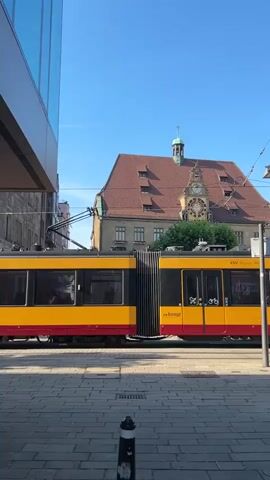 The media file shows a side view of a yellow and red tram passing by a historic building with a red tiled roof and a clock tower on a sunny day. The tram is in motion, indicated by the blur of its wheels and the slight bending of its articulated sections. Two trams are visible, one slightly in front of the other, with their front ends nearly touching.

The building behind the tram has a distinctive Mansard roof with multiple dormer windows and a central cupola. The clock on the tower is visible and appears to be functioning. Green trees are planted in front of the building, adding to the urban park-like feel. The scene is set on a paved street, with a sidewalk in the foreground. A street lamp and overhead wires for the tram are also visible.

The visible text on the tram includes "KVV. Bewegt alle." which is a slogan for the Karlsruher Verkehrsverbund (KVV), a public transport association in the Karlsruhe region of Germany. There are also symbols indicating accessibility for people with disabilities and bicycles. Another piece of text on the tram appears to be a series of numbers, possibly a vehicle identification number.

The sky is clear and blue, suggesting a pleasant, sunny day. The shadows cast by the tram and the building indicate it is likely mid-morning or early afternoon. The overall impression is one of a typical urban scene in a German city, with public transportation playing a central role. There are no clearly identifiable people in the foreground or on the tram, but a few figures can be seen through the tram windows. The atmosphere is calm and orderly, reflecting the efficient nature of public transport.