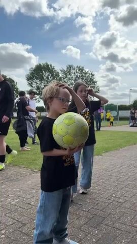 Two children, a boy and a girl, stand outdoors on a paved surface under a partly cloudy sky. The boy, on the left, has blond hair and glasses and holds a worn, yellow and black soccer ball in front of him with both hands. He wears a black t-shirt with yellow text and blue jeans, looking towards his right. The girl stands slightly behind and to the right of the boy, possessing long dark hair and wearing an identical black t-shirt with yellow text, including "Aufstieg," and blue jeans. She is touching her face or hair with her right hand while looking to her right. In the background, a grassy field with a soccer goal is visible, along with several adults, some in dark attire, and other individuals further in the distance.