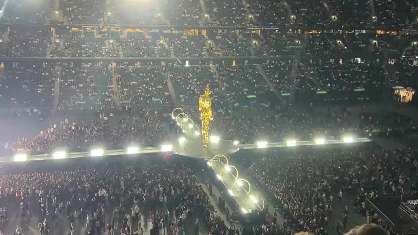 The video captures a wide-angle view of a large indoor stadium filled with a dense crowd of people, most of whom are standing. At the center of the stadium, a brightly lit stage is set up with a massive, golden, humanoid statue standing tall on a platform. The statue is illuminated by spotlights, creating a strong glow.

Leading away from the statue, a runway-like pathway is lined with a series of illuminated, circular arches. The stadium seating areas are visible in the background, also filled with spectators. Smoke or fog drifts across the stage and into the crowd, adding a dramatic effect. The lighting is predominantly bright and stage-like, with some areas of shadow in the upper tiers of the stadium. The overall impression is that of a large-scale event, possibly a concert, awards ceremony, or performance.
