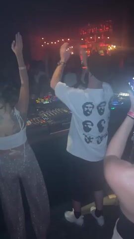 A group of people are at a DJ booth at a nightclub. The DJ, a man wearing a white t-shirt with a graphic print and a dark hat, is standing behind the DJ equipment, facing away from the camera, with his hands raised in the air. To the left of the DJ, a woman with curly brown hair, wearing a white top and patterned pants, is also facing away from the camera with her left arm raised. Other people are visible in the background, including a man in a white shirt standing behind the DJ. The environment appears to be dark, with red lighting and string lights visible in the background, suggesting a nightlife setting.
