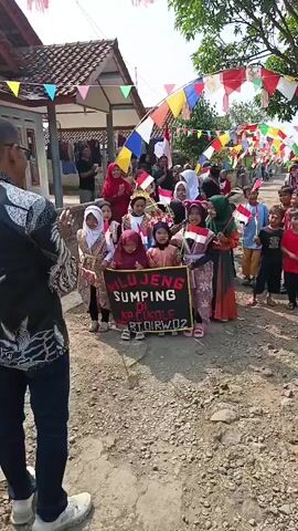 A group of children, many wearing traditional clothing and headscarves, are participating in a parade. Some children are holding small Indonesian flags, and a few are holding a banner that reads "WILUJENG SUMPING KP CIKOLE RT DIRW02". The children are walking on a dirt path lined with houses decorated with colorful triangular flags and streamers. Adults are standing on the sides, watching and some are clapping. The atmosphere appears festive.