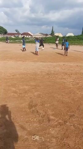 A group of children are playing soccer on a dirt field under a cloudy sky. There are several houses and trees in the background. One child is kicking the soccer ball while others are running and watching.