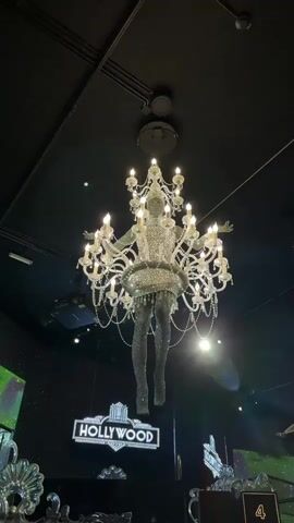 The video shows a low-angle shot of a chandelier in a dimly lit room. The chandelier is shaped like a female figure adorned with crystals, with a skirt and leggings. The figure appears to be suspended from the ceiling, with its arms outstretched. The chandelier has multiple arms, each holding a candle-shaped light bulb, and is decorated with hanging crystals. Below the chandelier, a sign with the word "HOLLYWOOD" in illuminated letters is visible. The background is dark, with some areas of sparkle and faint green light on the left side. The ceiling is black with visible conduits and fixtures. The overall impression is of a theatrical or glamorous setting.