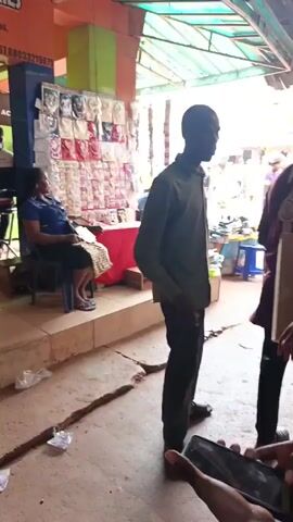 A person is filming while standing in a covered marketplace. Several people are walking around. A woman sits on a step near a stand selling small items and a man sits on a stool nearby. Another man sits on the ground near a table laden with items, likely shoes and sandals. The floor of the marketplace is concrete with cracks. There are additional vendors and shoppers further down the passageway.