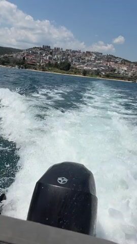 The video begins with a close-up shot of the rear of a boat, showing the outboard motor creating a large wake. The camera then pans out to reveal a coastal town with buildings on a hillside, and the sea. Two large passenger ferries are docked in the distance. The sky is clear and blue, indicating a sunny day. The boat appears to be moving at a moderate speed, generating significant white water.
