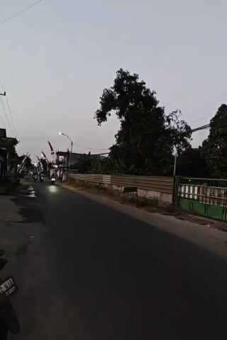 The video shows a street scene at dusk. The camera pans from left to right, capturing a road with some traffic. On the left side of the road, there are flags and a few motorcycles. Further down the road, a building with a sign that reads "WARKOP 49 JOGO ROSO" is visible. There are tables and benches outside the building, suggesting it is a cafe or eatery. The sky is overcast, and the lighting suggests it is either early morning or late evening. Several power lines are visible overhead. A motorcycle with a rider passes by in the foreground.
