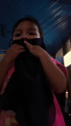 A young child wearing a bright pink shirt stands in front of a blue tarp ceiling. The child is holding a black piece of fabric, possibly a mask or scarf, up to their face, covering their nose and mouth. Their eyes are visible and they appear to be looking towards the camera with a playful or shy expression. The background is dimly lit, with some wooden structures and a bright light source visible on the right side of the frame. The child's hands are gripping the fabric, and their fingers are visible as they adjust it over their face. The child's hair is dark and appears to be pulled back from their face.