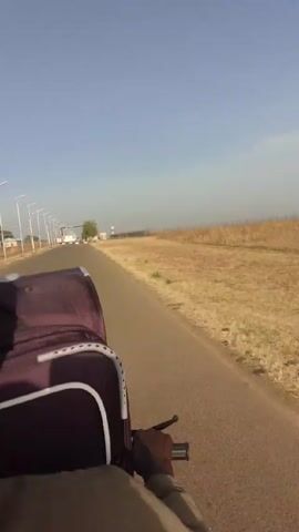 The video shows a first-person perspective from someone riding a motorcycle. In front of the rider is a purple suitcase strapped to the back of the bike. The rider is wearing a pink and beige knit hat and a tan jacket. The motorcycle is traveling down a paved road lined with streetlights. There is dry, grassy land on either side of the road. In the distance, there is a gate with red and white barriers. The sky is blue and clear.