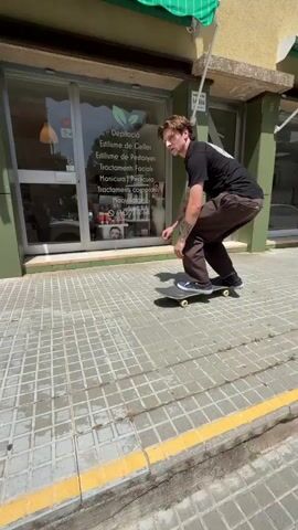 A young man in a black t-shirt and brown pants is skateboarding. He begins by riding down a sidewalk and then attempts to grind on a metal railing, but he falls off his skateboard. The skateboard rolls away from him down a ramp. The background shows a building with shops.