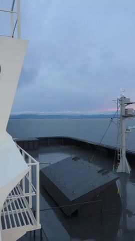 The video shows a view from the deck of a ship. The sky is overcast with some streaks of pink and orange indicating either sunrise or sunset. The sea is calm and stretches to the horizon where mountains are visible. On the deck, there is a large, dark, rectangular object with an angled top, secured by ropes. To the right, a tall, white pole with various instruments, including what appears to be a wind anemometer, is mounted. The deck itself is wet and reflective, suggesting recent rain or sea spray. In the foreground, a white railing and stairs are visible on the left side of the frame. The overall impression is one of a serene maritime scene at dawn or dusk.