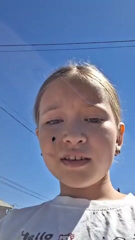 A young girl with blonde hair is filmed from the chest up, looking into the camera. She is wearing a white t-shirt with a cartoon design. She has a dark spot on her right cheek, possibly a mole or a smudge of dirt. The background shows a clear blue sky, power lines, a pale pink house with a white roof, and some greenery. The girl appears to be outside on a sunny day.