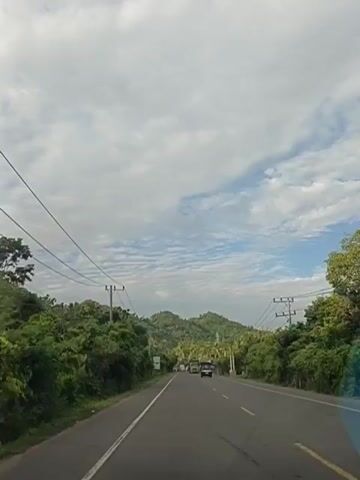 The video shows a straight asphalt road with a white line on the left and a yellow line on the right. The road is bordered by lush green trees and vegetation on both sides. Overhead power lines are visible, supported by wooden poles. In the distance, rolling green hills are present under a cloudy sky with patches of blue. There are several vehicles on the road. A dark-colored SUV is in the right lane, and a green truck is in the left lane, passing a few cars. The truck has a license plate visible. The time stamp at the beginning of the video indicates it starts at 00:00.