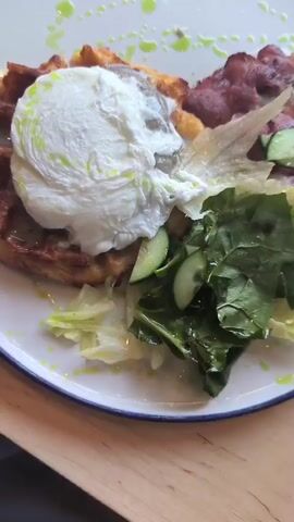 A close-up, overhead, static shot shows a plate of food. The plate is white with a blue rim. On the plate, a golden-brown waffle is topped with a perfectly poached egg. The egg is fluffy and white with a slight drizzle of green sauce. To the side of the waffle, there is a salad consisting of mixed greens and sliced cucumbers. Several strips of crispy bacon are arranged behind the waffle. The entire dish is lightly garnished with a green drizzle of sauce. The plate is set on a light-colored wooden surface.
