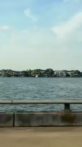 The video shows a body of water from a bridge. A row of houses can be seen across the water. The sky is blue with wispy clouds. The bridge is made of concrete with a metal railing. The video appears to be shot from a moving vehicle.