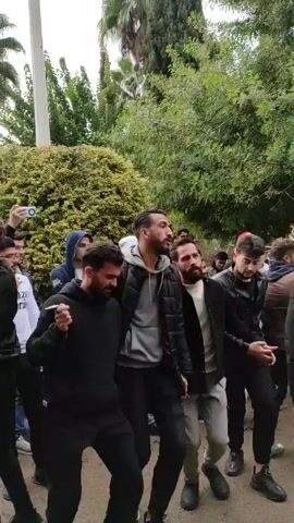 Several men dance in an outdoor setting. The group is diverse in appearance, wearing jackets and jeans. Some sing along to music that can be heard in the background. They move in a way that looks joyful. The area surrounding them has trees and greenery. The weather appears to be overcast or cloudy.