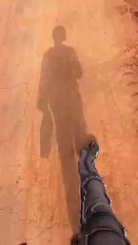 A first-person perspective shot shows someone walking on an orange, cracked surface, likely a dirt road or path. The viewer can see the person's lower legs, clad in unique denim pants with patches, and their feet in dark sandals. The sun casts a distinct shadow of the person onto the ground ahead, suggesting they are wearing a longer shirt or jacket. The surrounding area seems to be arid or contain reddish soil.