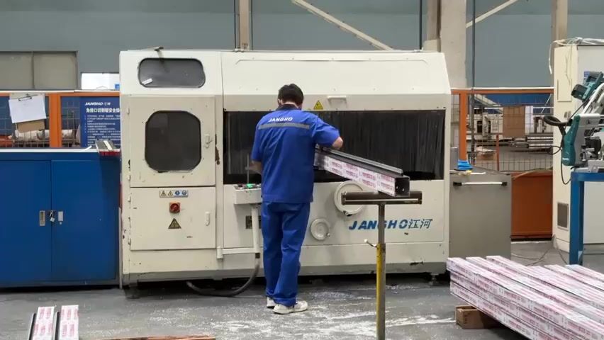 A worker in a blue uniform and a face mask stands in front of a large white machine with the word “JANGHO 江河” printed on its front. He carefully positions a long metal bar onto a support attached to the machine. The machine activates, generating sparks and a spray of coolant as it cuts the metal. To the worker's right, a stack of similar metal bars rests on the floor. Behind him, another worker walks by. The environment appears to be a factory floor, with various pieces of equipment and materials scattered around.