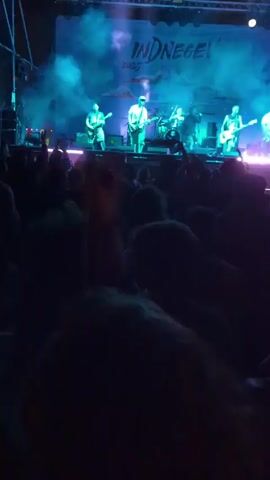 A band is performing on an outdoor stage at a music festival, with a large crowd in front of the stage. The band consists of six members, including a drummer and several guitarists. The stage is lit with blue and purple lights, and smoke effects are being used. The band's name, "INDNEGEV 2023," is displayed on a banner behind them. The crowd is mostly silhouetted, but they are clearly enjoying the performance. Some people are raising their hands in the air. The environment appears to be outdoors at night.