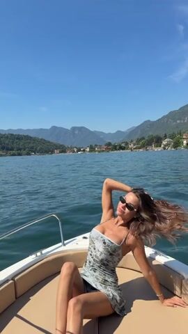 A woman wearing sunglasses and a light-colored dress with a floral print is sitting on the edge of a boat. The boat is on a large body of water, likely a lake, with mountains and trees visible in the background. The sky is clear and blue. The woman is posing with her hand on her head and the other resting on her leg. The boat appears to be moving, as indicated by the slight blur of her hair.