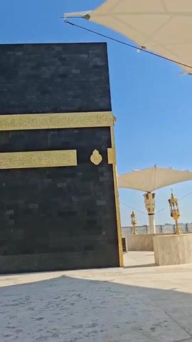 The video begins with a low-angle shot of a replica of the Kaaba in Mecca, its black stone exterior adorned with gold calligraphy and a gold-embossed emblem. The Kaaba is under a clear blue sky, with a large white canopy structure casting a shadow on the ground. A golden lantern-like sculpture stands in front of the Kaaba. The camera pans to the right, revealing more of the surrounding area.

As the camera continues to pan, the Kaaba replica moves out of frame, replaced by a white awning supported by ornate pillars. A palm tree is visible in the background, along with a building. A black van is parked in the foreground, and further in the distance, a silver car is seen driving. A person is standing near the palm tree. The sky remains a vibrant blue with scattered white clouds. The video captures a bright, sunny day in an outdoor setting that appears to be a tourist attraction or religious site. The overall impression is one of reverence and peacefulness.