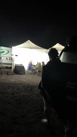 At night, two people stand at the order window of a lit-up food truck while a worker stands inside. The truck is black, with the words “Aquí Empieza lo Bueno” written on the side. The truck has a sign on top that is round and illuminated. A white tent is visible behind the truck, and a person walks in front of the truck.