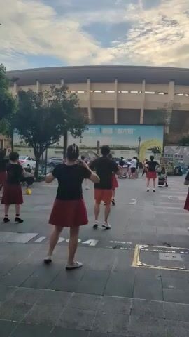 A group of people are dancing outdoors in front of a building that appears to be a stadium. They are wearing matching black tops and red skirts or shorts. The sky is cloudy and the lighting suggests it is either early morning or late afternoon. The ground is paved with square tiles.