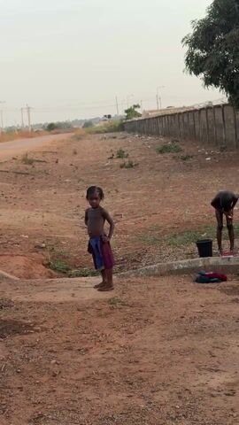The video shows children in an outdoor setting. Initially, a young girl stands with her arms at her sides, while another individual, possibly a child, is bent over near a bucket. As the camera pans, two more children come into view, one sitting wrapped in a purple cloth and another sitting beside them. Subsequently, a child holding a stick stands near a pot on a stove while an adult figure bends over in the background. The environment appears to be a rural or semi-rural area with a dirt ground, a low wall, and some vegetation. The children are partially clothed, and appear to be involved in washing or preparing for a meal.