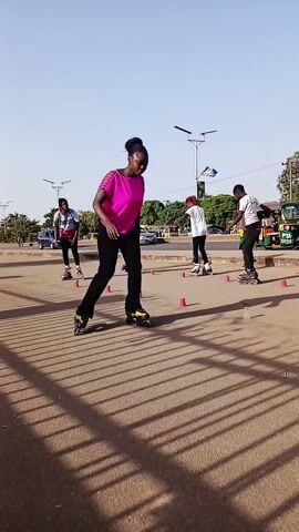 The video shows a woman wearing a bright pink short-sleeved shirt and black pants as she rollerblades between small orange cones. She is the primary focus. Her hair is up in a bun. Other individuals on rollerblades are nearby. Some of them are skating and some are standing. Some wear white shirts. The scene takes place on a paved area that appears to be part of a road. There are shadows from a metal fence and trees cast on the ground. There are several vehicles and buildings visible in the background. The sky is clear and blue. A billboard is visible in the background on the right side of the frame. Overall, the setting appears to be an urban outdoor area.